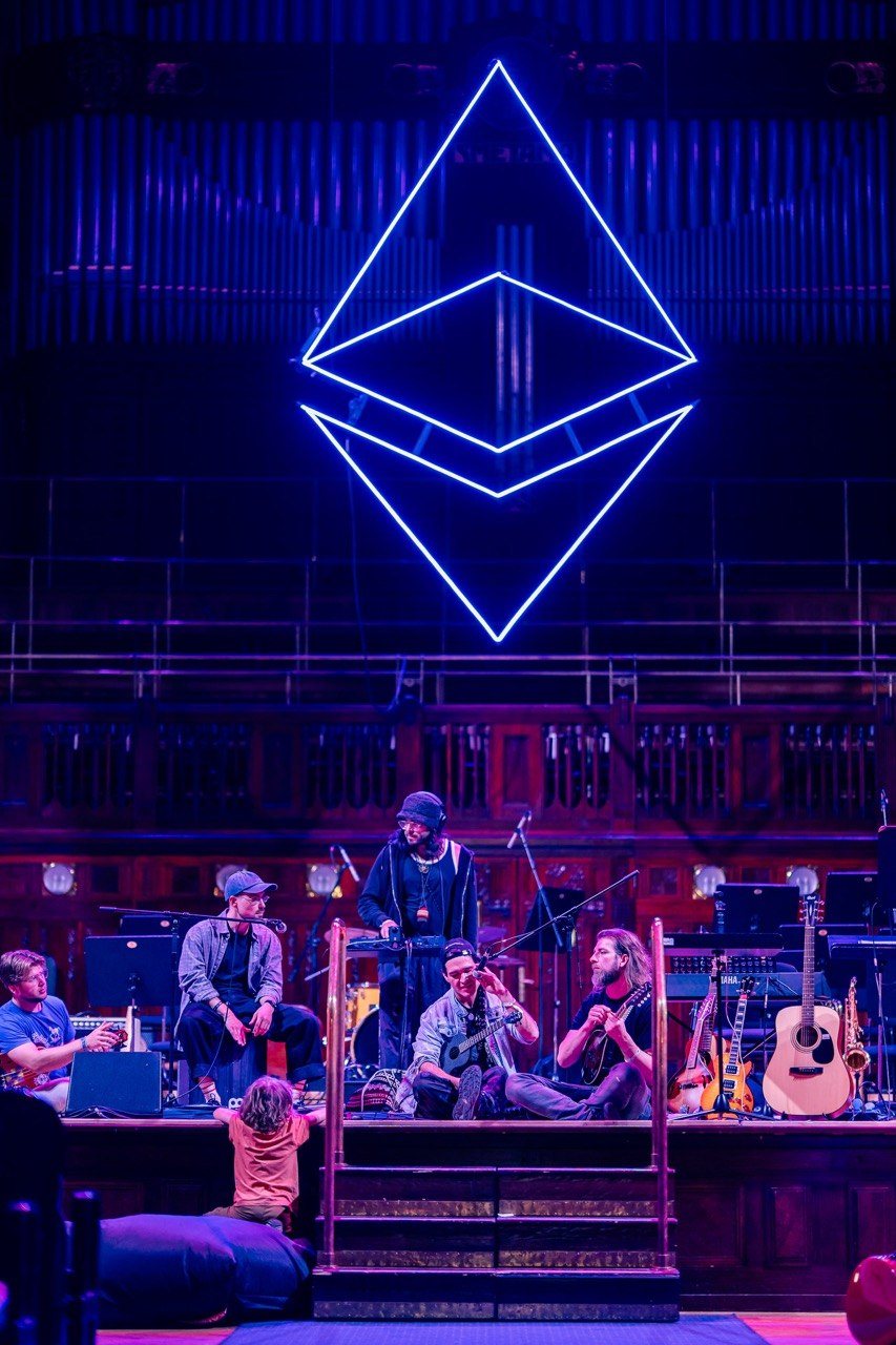 The Ethereum sigil in neon above the stage, musicians in a circle below, a child reaching up